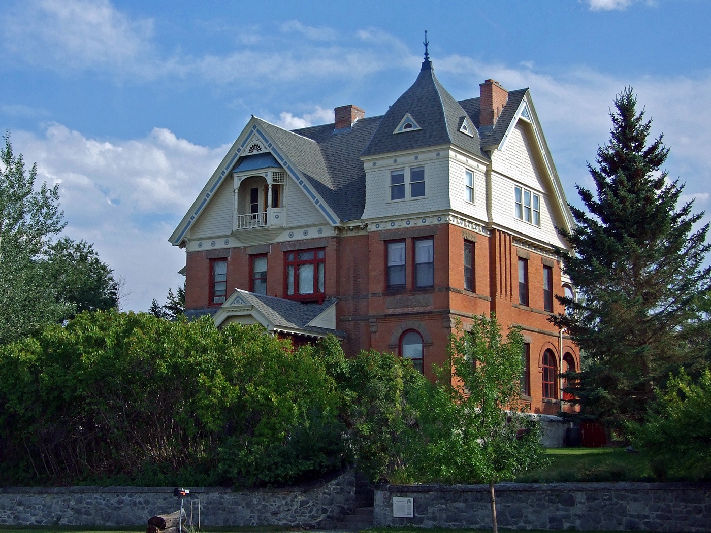 The T. H. Kleinschmidt House at 1823 Highland Avenue, Helena, Montana: a three-storey Queen Anne residence of red brick below and pale shingle above, with a steep roof, a corner turret, a recessed second-floor balcony and tall brick chimneys, photographed in summer daylight behind a stone retaining wall