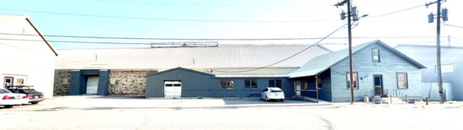 The full street elevation of 1316 Bozeman Avenue today: the exposed stone bay with its loading door at left, the long metal-clad warehouse wall, and the clapboard office wing with its porch at right