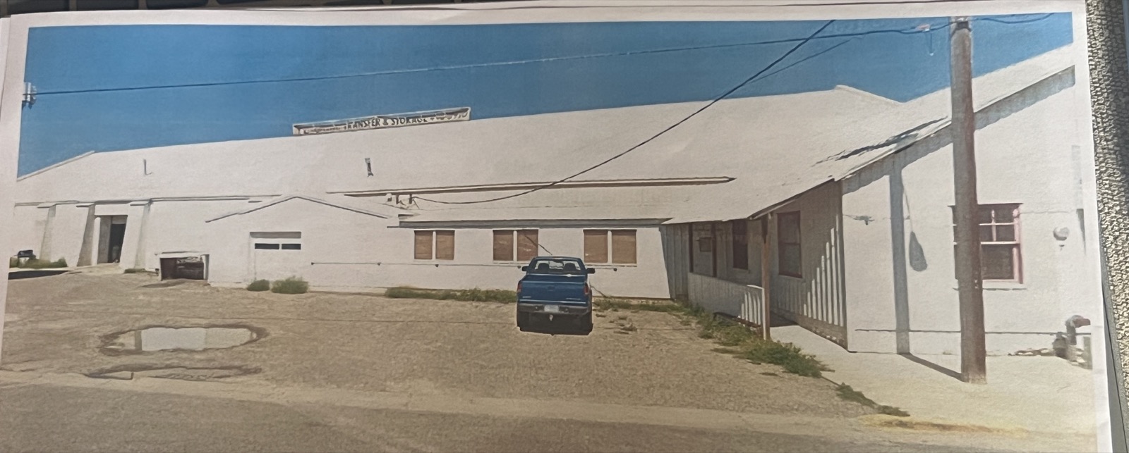 A photographic print of the warehouse at 1316 Bozeman Avenue in the Capital Transfer & Storage years: the long building painted white, the words Transfer & Storage across the upper wall, a blue pickup parked in front, the office wing at right